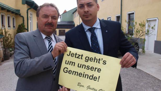 Zwei Männer halten ein Schild mit der Aufschrift „Jetzt geht's um unsere Gemeinde! Bgm. Gustav Glöckler“.