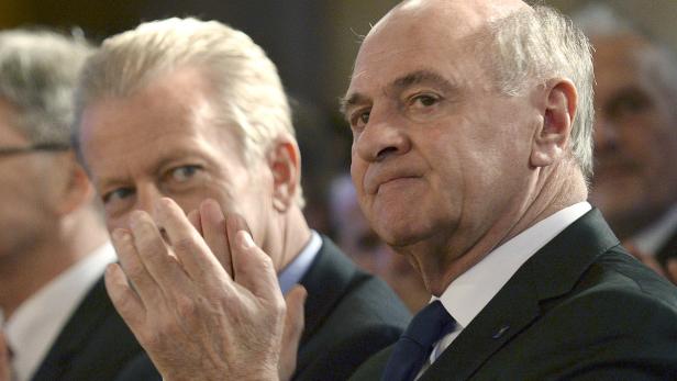 Zwei ältere Männer in Anzügen, einer mit der Hand am Gesicht.