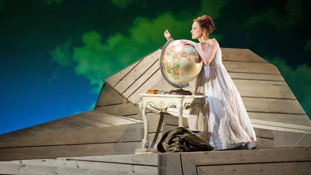 Eine Frau in einem weißen Kleid steht auf einer Bühne und betrachtet einen Globus.