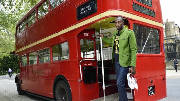 Usain Bolt steht vor einem roten Doppeldeckerbus und hält ein Paar Schuhe in der Hand.