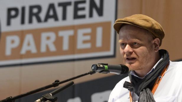 Ein Mann mit Schiebermütze spricht am Mikrofon vor einem Banner der Piratenpartei.