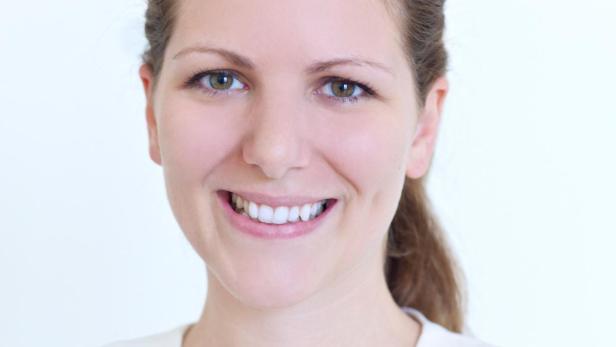 Eine lächelnde Frau mit braunen Haaren und einem weißen Blazer vor einem weißen Hintergrund.