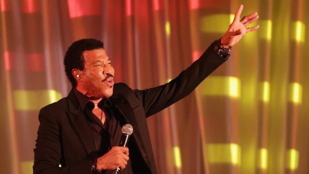 Lionel Richie tritt in einem schwarzen Anzug mit einem Mikrofon in der Hand auf einer Bühne auf.