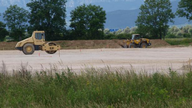 Eine Walze und ein Bagger stehen auf einer Baustelle vor einer Hügellandschaft.