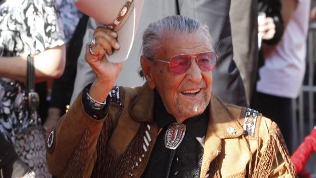 Ein älterer Mann mit rosa Sonnenbrille und Cowboyhut lächelt.