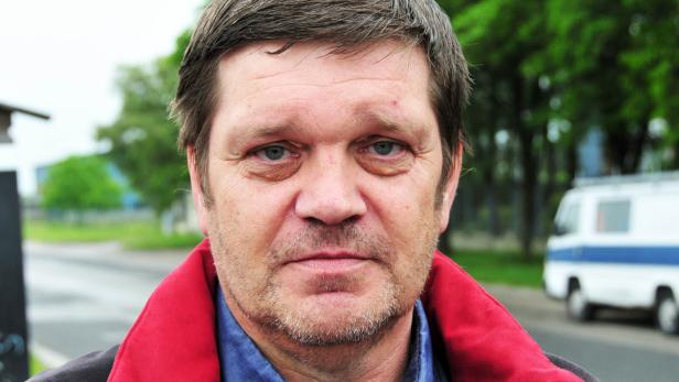 Porträt eines Mannes mit Bart und rotem Kragen vor unscharfem Hintergrund.