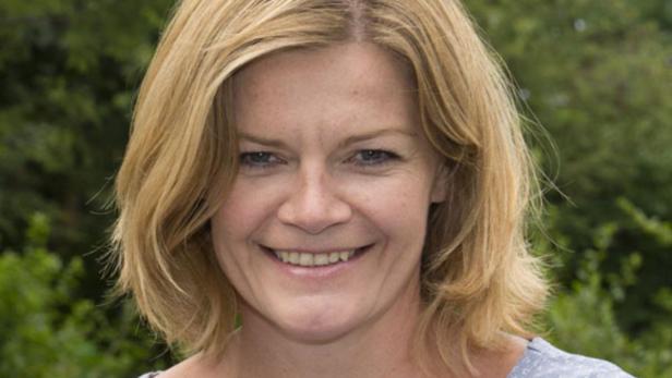 Eine lächelnde Frau mit blonden, schulterlangen Haaren vor grünem Hintergrund.