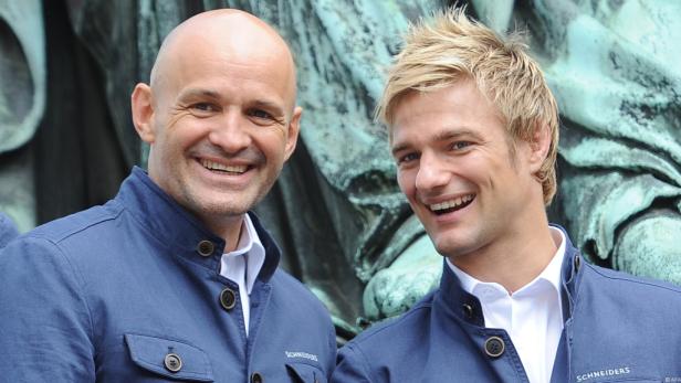 Zwei Männer lächeln in blauen Jacken vor einem Denkmal in die Kamera.