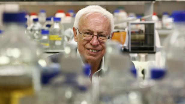 Ein lächelnder älterer Mann mit Brille steht in einem Labor hinter vielen Flaschen.