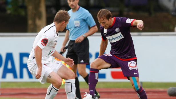 Zwei Fußballspieler stehen sich auf dem Spielfeld gegenüber, ein Schiedsrichter im Hintergrund.