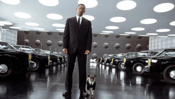 Will Smith steht mit einem Mops im Anzug vor einer Reihe schwarzer Autos.