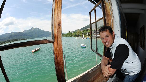 Ein Mann lehnt an einem Fenster mit Blick auf einen See mit Bergen im Hintergrund.