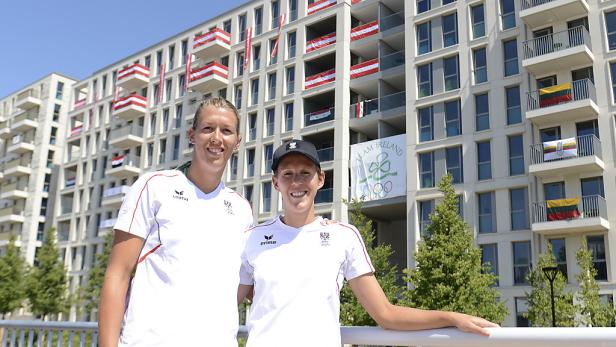 Zwei Sportlerinnen des österreichischen Teams posieren vor einem Gebäude mit Flaggen verschiedener Nationen.