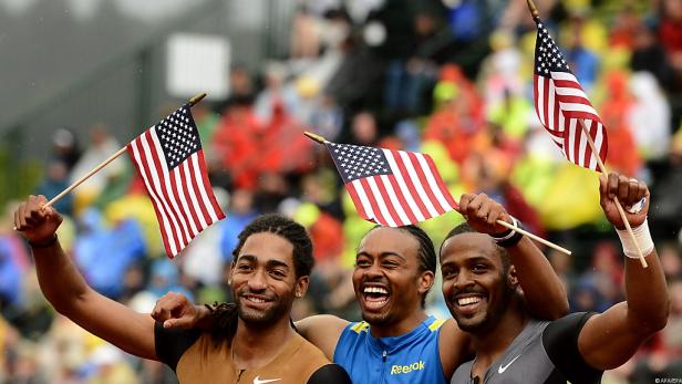 Drei lachende Athleten schwenken US-amerikanische Flaggen.