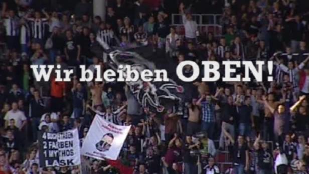 Eine Menschenmenge jubelt auf einer Tribüne mit Bannern und der Aufschrift „Wir bleiben OBEN!“.