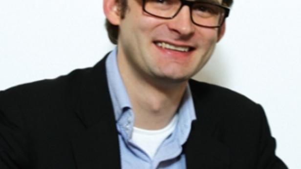 Ein lächelnder Mann mit Brille und dunklem Sakko vor einem weißen Hintergrund.