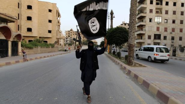 Eine Person mit einer IS-Flagge und einem Gewehr geht eine Straße entlang.