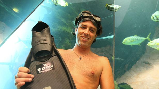 Ein Mann mit Taucherbrille hält eine Schwimmflosse vor einem Aquarium mit Fischen.