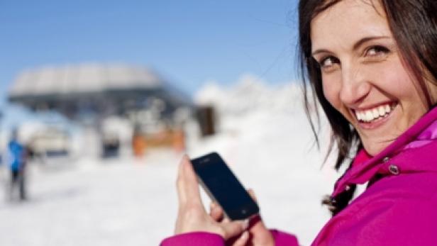 Eine lächelnde Frau mit Smartphone vor einer verschneiten Berglandschaft.