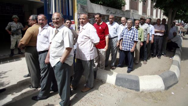 Eine Menschenmenge steht vor einem Gebäude Schlange, bewacht von einem Soldaten.