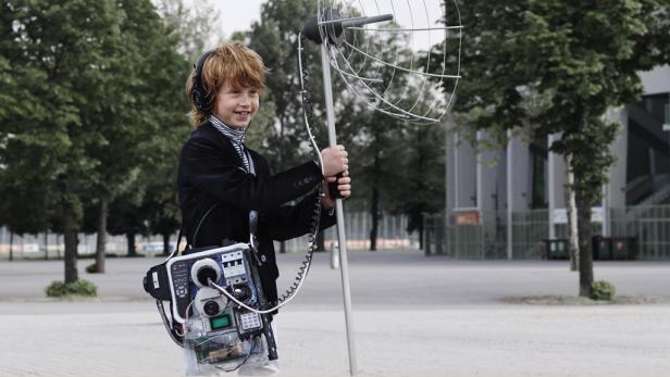 Ein Junge mit Kopfhörern und einer selbstgebauten Apparatur mit Antenne steht im Freien.