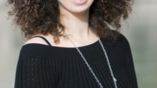 Eine lächelnde Frau mit Brille und lockigem Haar trägt eine lange Halskette.