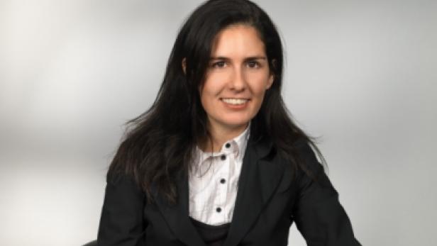 Eine Frau mit dunklen Haaren lächelt in einem schwarzen Blazer.