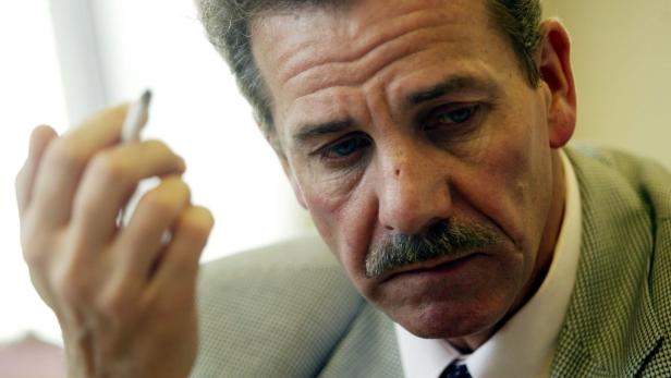 Ein Mann mit Schnurrbart hält einen Stift in der Hand und wirkt nachdenklich.