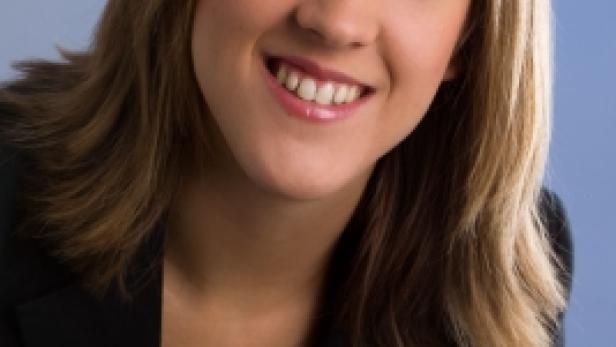 Eine lächelnde Frau mit blonden Haaren und einem schwarzen Blazer vor blauem Hintergrund.