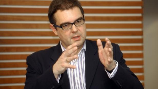 Ein Mann mit Brille und Anzug gestikuliert vor einer Holzwand.