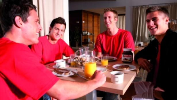 Vier junge Männer sitzen an einem Tisch und frühstücken mit Nutella und Orangensaft.