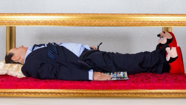 Eine Wachsfigur von Silvio Berlusconi liegt in einem gläsernen Sarg mit Mickey-Mouse-Hausschuhen.