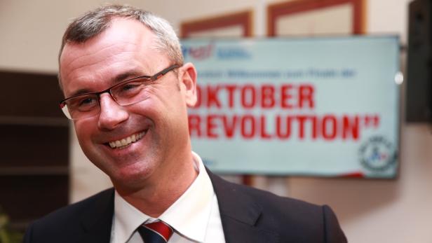 Ein lächelnder Mann mit Brille und Anzug vor einem unscharfen Hintergrund mit der Aufschrift „Oktober Revolution“.