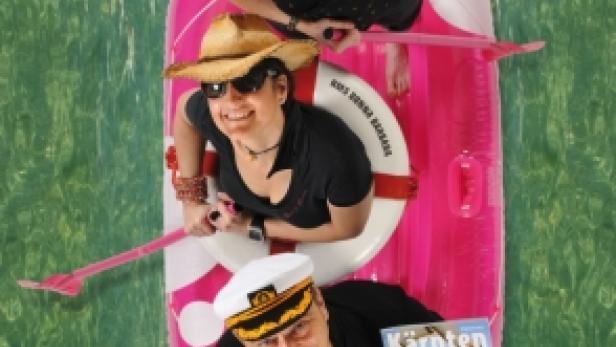Drei Personen in einem aufblasbaren Boot mit einem „Kärnten“-Magazin vor grünem Hintergrund.