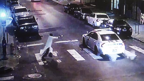 Auf einer dunklen Straße zielt eine Person mit einer Waffe auf ein Polizeiauto.