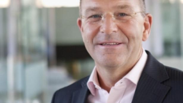 Ein lächelnder Mann mit Brille und Anzug vor einem Bürogebäude.