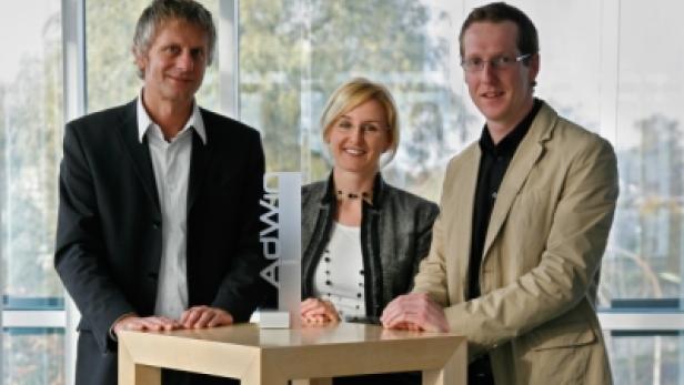 Drei Personen posieren mit einem AdWin-Award an einem Tisch.