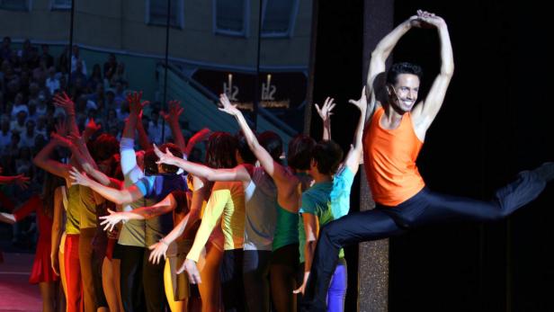 Ein Tänzer springt vor einer Gruppe anderer Tänzer auf einer Bühne.
