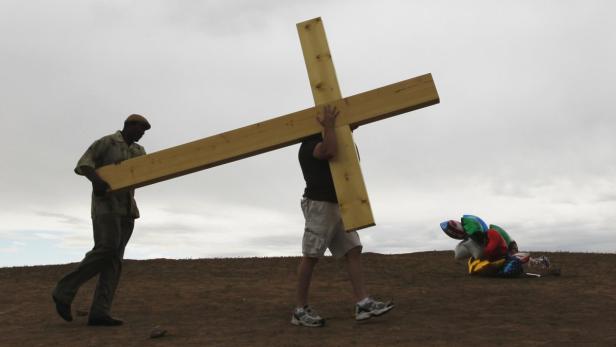 Zwei Männer tragen ein Holzkreuz auf einem Hügel, im Hintergrund Ballons.