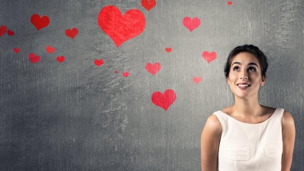 Eine junge Frau lächelt verträumt vor einer Wand mit roten Herzen.