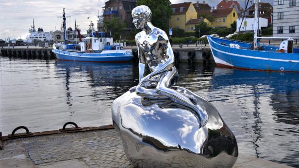 Eine glänzende, silberne Meerjungfrau-Skulptur sitzt an einem Hafen.