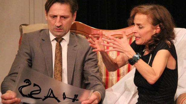 Eine Frau gestikuliert aufgeregt, während ein Mann mit einem beschrifteten Blatt verunsichert daneben sitzt.