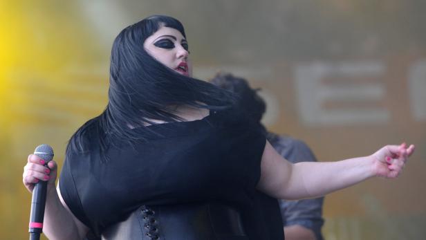 Eine Sängerin mit dunklem Haar und auffälligem Make-up tritt mit einem Mikrofon auf einer Bühne auf.