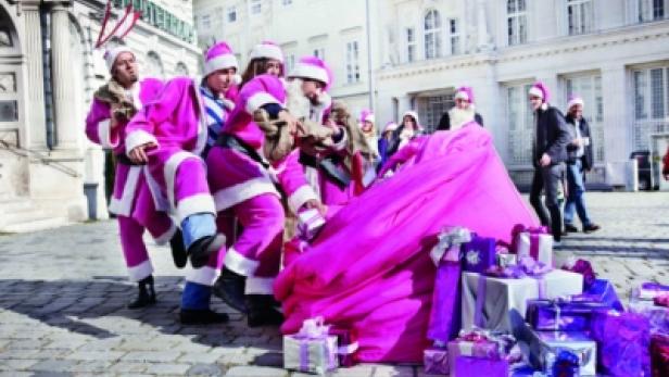 Eine Gruppe von Personen in pinkfarbenen Weihnachtsmannkostümen trägt einen Sack voller Geschenke.