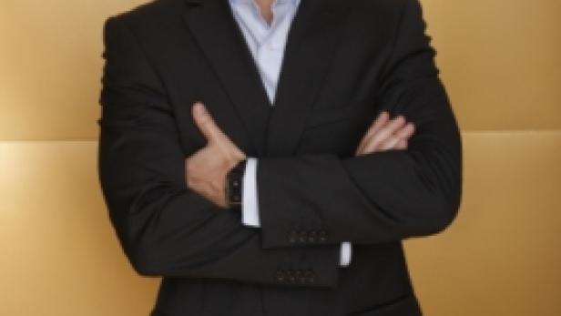 Ein Mann mit Brille und Anzug steht mit verschränkten Armen vor einem goldenen Hintergrund.