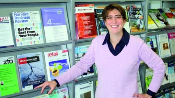Eine Frau steht lächelnd vor einem Regal mit Zeitschriften in einer Bibliothek.