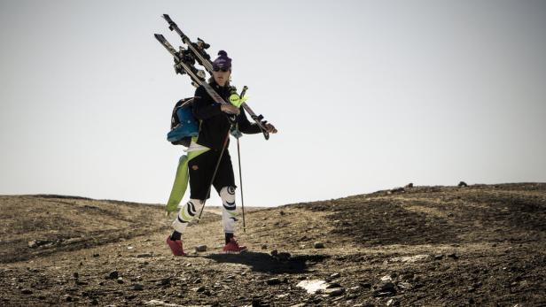 Eine Frau trägt Skier und Skischuhe auf einem felsigen Hügel.