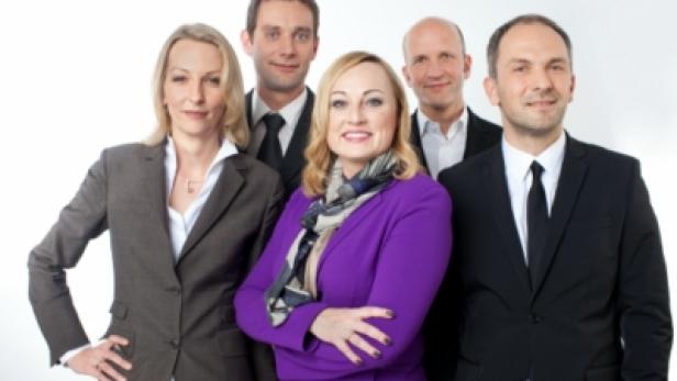 Eine Gruppe von fünf Personen in formeller Kleidung posiert vor einem weißen Hintergrund.