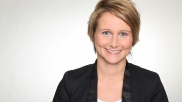 Eine lächelnde Frau mit blonden Haaren und einem schwarzen Blazer vor einem hellen Hintergrund.