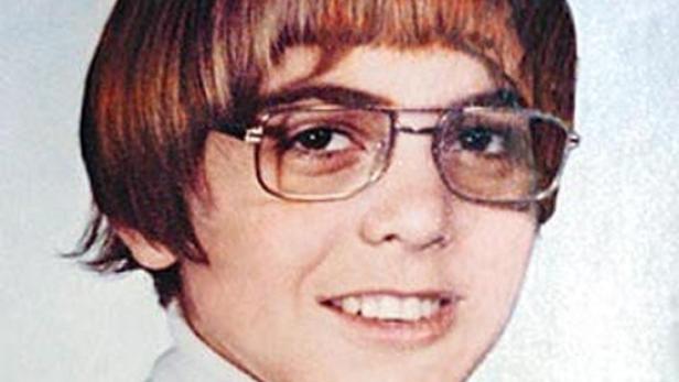 Ein junger George Clooney mit braunen Haaren, Pony und Brille lächelt.